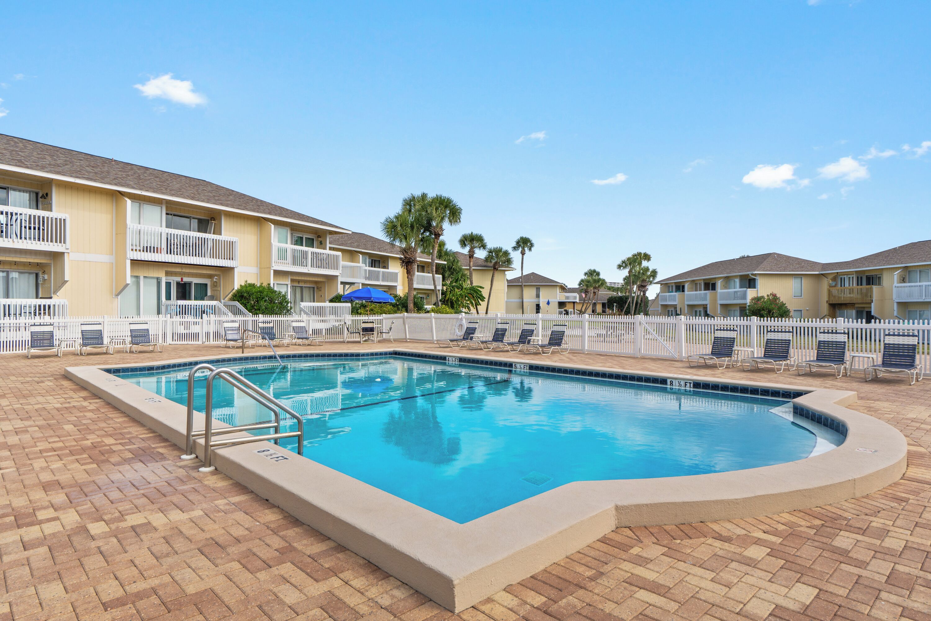 A swimming pool with a brick deck, surrounded by a white fence and lounge chairs.