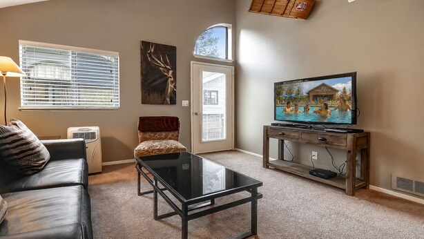 Una sala de estar con un sofá, una mesa de centro de vidrio, un televisor sobre un mueble de madera, una chimenea y una puerta que da al exterior.