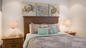 A bedroom with a wooden headboard, a bed with a floral-patterned bedspread, multiple pillows, two bedside tables with lamps, and three wall-mounted artworks depicting sea creatures. - Direct Ocean & Boardwalk Views - Beautifully Appointed Condo - Parking & Wi-Fi (Hollywood)