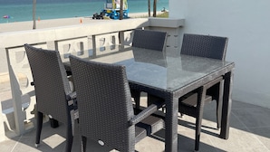 A patio with a glass-top table and wicker chairs, overlooking a beach with palm trees and a lifeguard tower. - Direct Ocean & Boardwalk Views - Beautifully Appointed Condo - Parking & Wi-Fi (Hollywood)