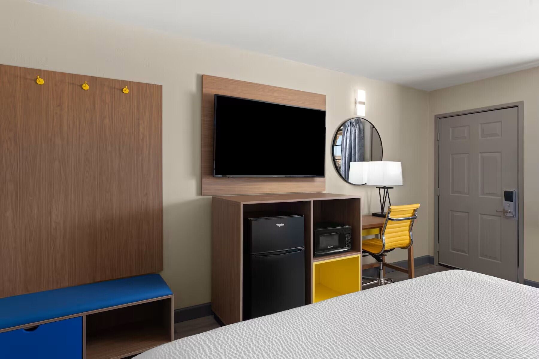 A hotel room with a bed, a TV mounted on the wall, a small refrigerator, a yellow chair, and a door.