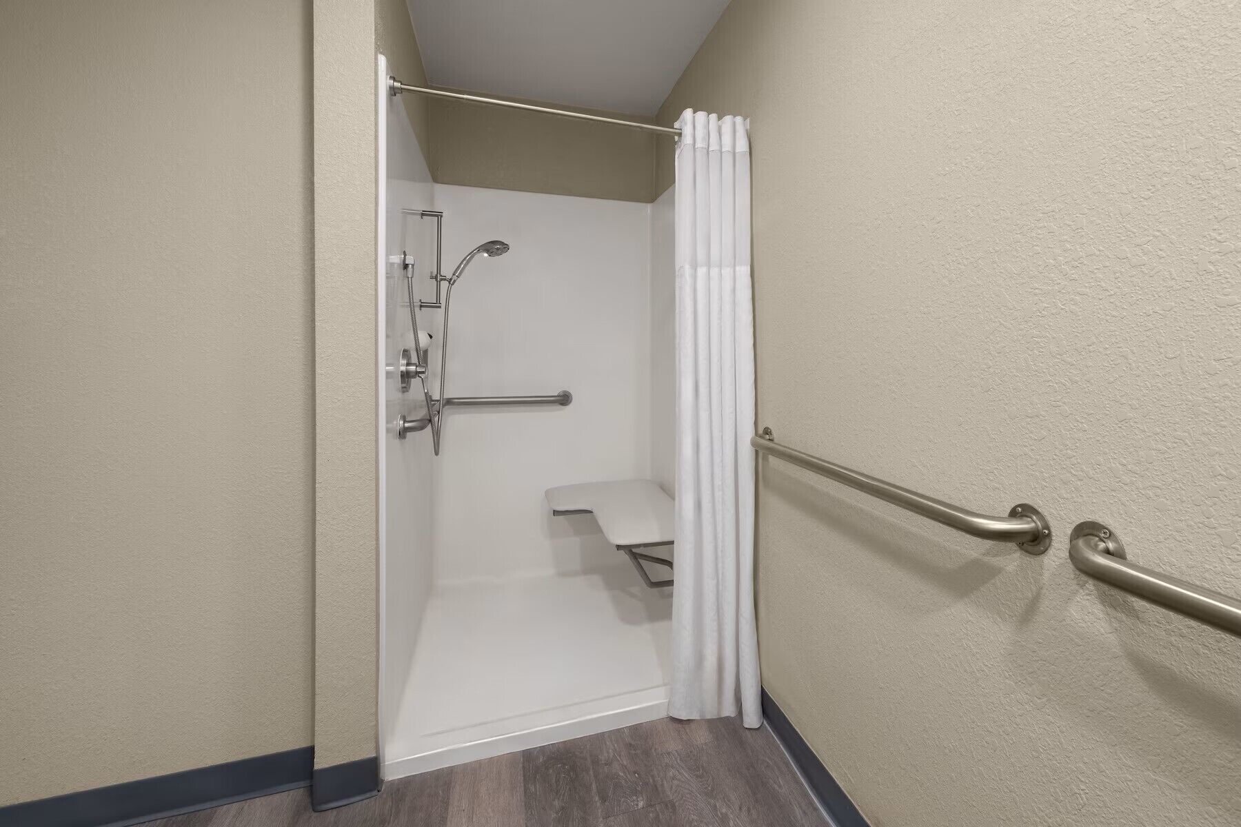 A shower area with a glass door, a showerhead, a handrail, and a white curtain.