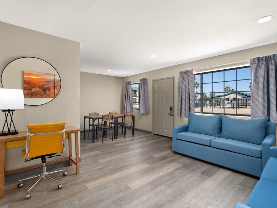 A modern hotel room with a blue sofa, a wooden desk with a chair, a dining table with chairs, and a large window with curtains.