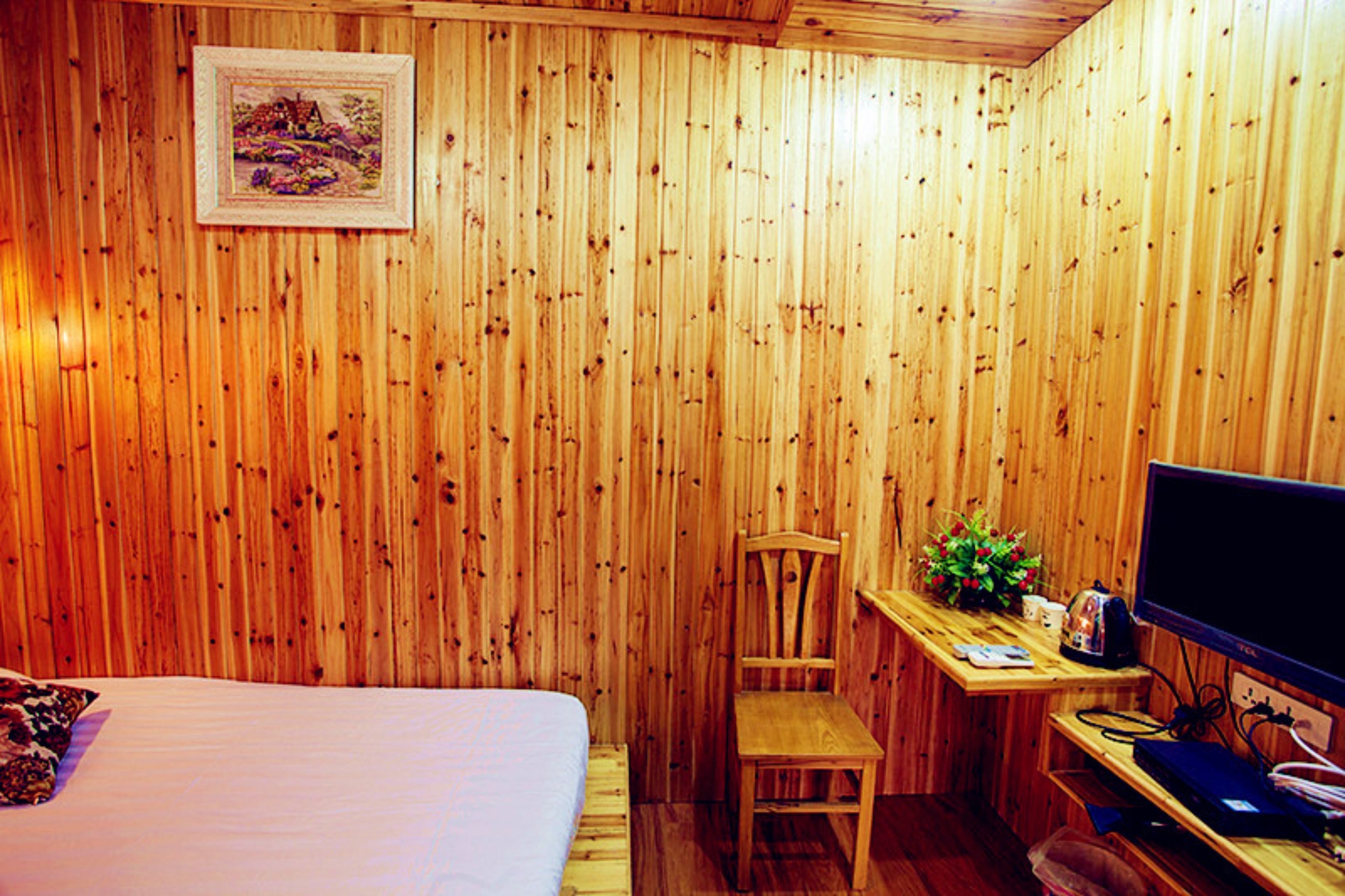 Ein Zimmer mit Holzwänden, einem Bett, einem Schreibtisch, einem Sessel, einem Fernseher und einem kleinen Tisch mit Pflanze und Tasse.