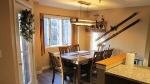 A dining area with a wooden table and chairs, a window with curtains, a hanging light fixture, and a staircase with skis.