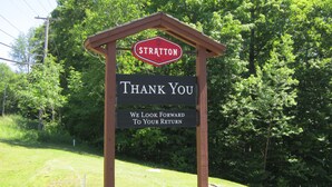 A wooden sign with a red and white logo, a black signboard with white text, and a backdrop of green trees.