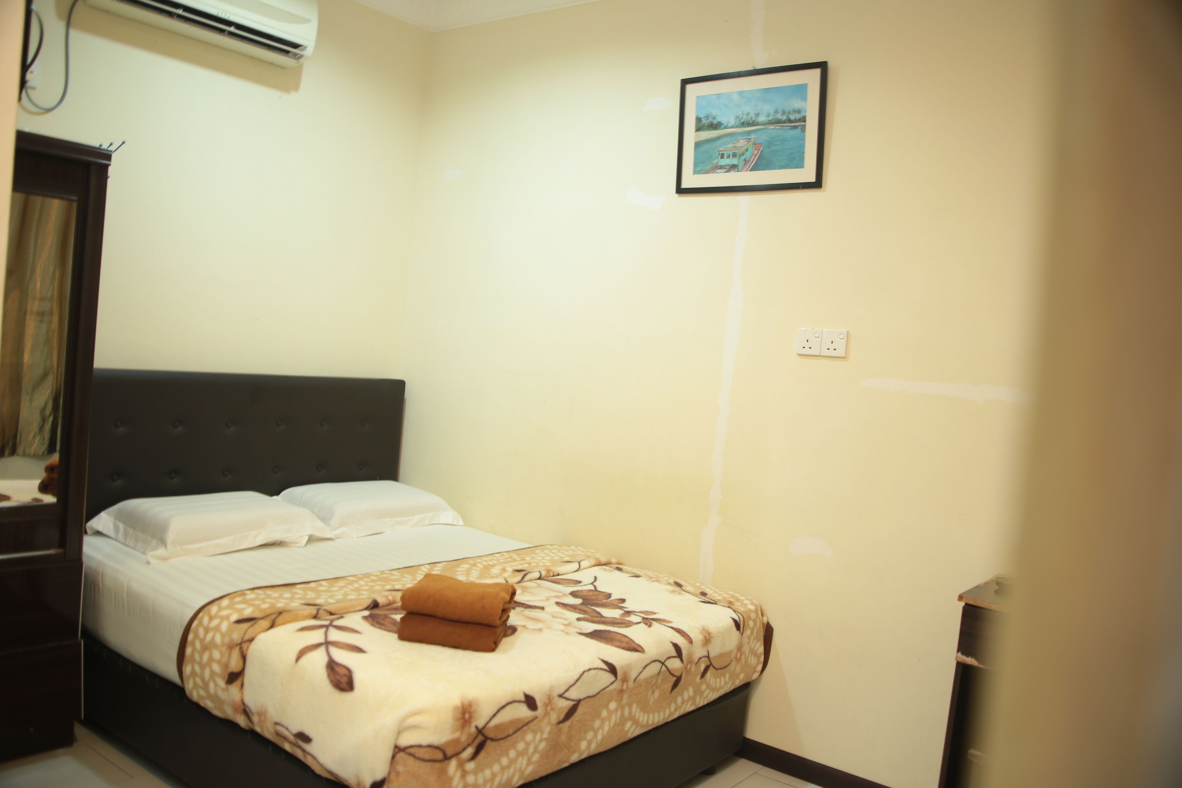A neatly made bed with a floral bedspread and a brown towel, a bedside table, a wall-mounted air conditioner, and a framed picture on the wall.