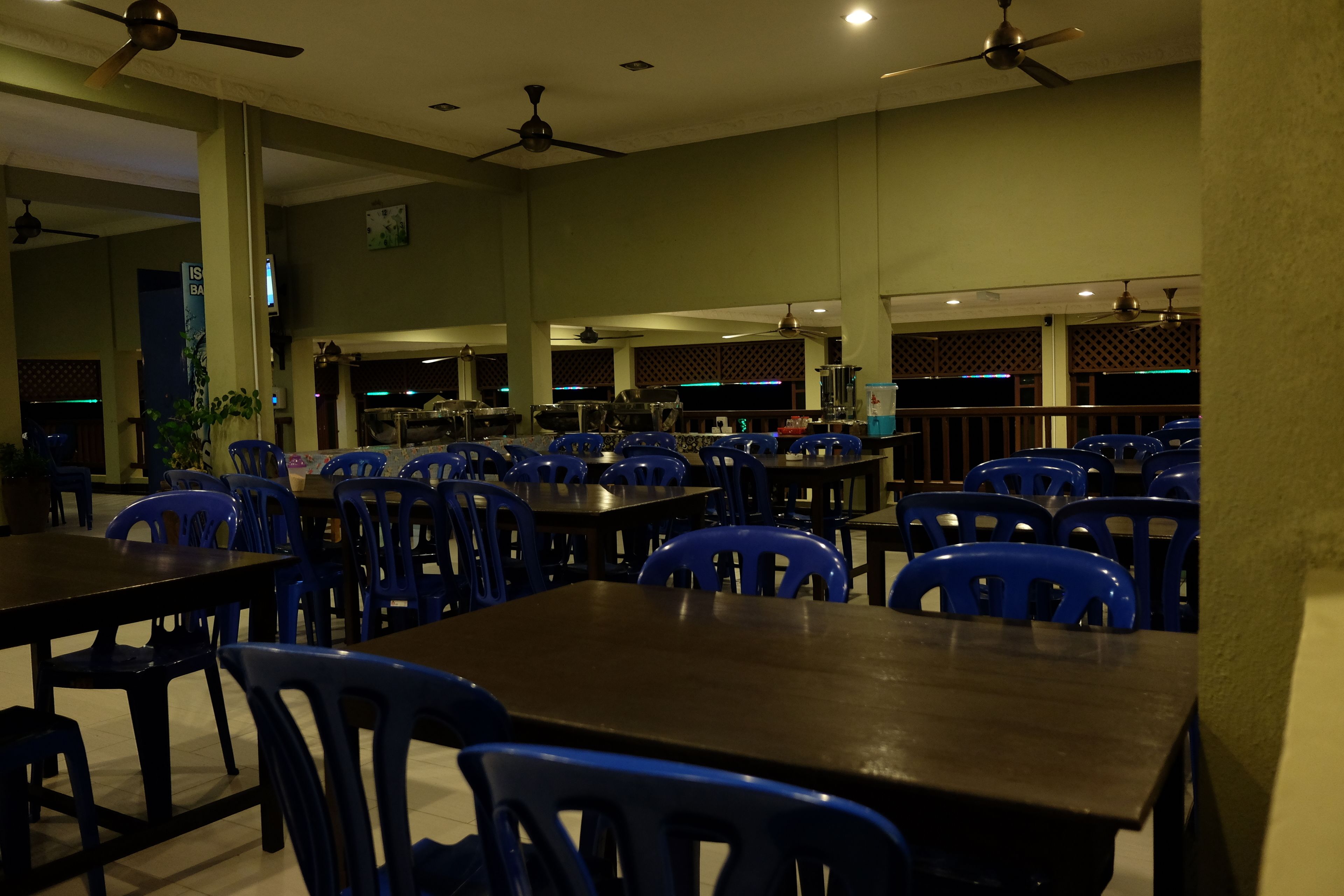 A spacious dining area with multiple tables and blue plastic chairs, ceiling fans, and a bar area in the background.