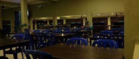A spacious dining area with multiple tables and blue plastic chairs, ceiling fans, and a bar area in the background.
