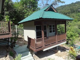 Una pequeña estructura de madera elevada con techo verde y un balcón, rodeada de vegetación natural y colinas.