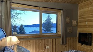 A cozy cabin room with a large window overlooking a snowy landscape, a fireplace, and a bed with pillows.