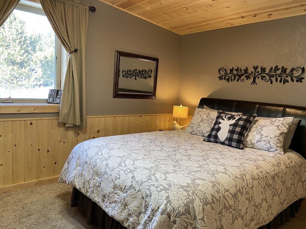 A bedroom with a large bed, a window with curtains, a framed picture on the wall, and a decorative wall piece.