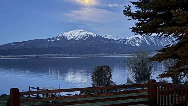 A deck with wooden chairs and a table overlooking a lake with mountains in the background.