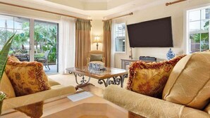 A living room with a glass coffee table, beige sofas, a flat-screen TV, and a view of greenery outside. - First Floor Condo, Near Pools and Attractions (Kissimmee)