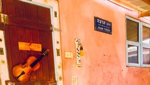 A pink building with a wooden door, a violin leaning against it, and a sign in Hebrew.