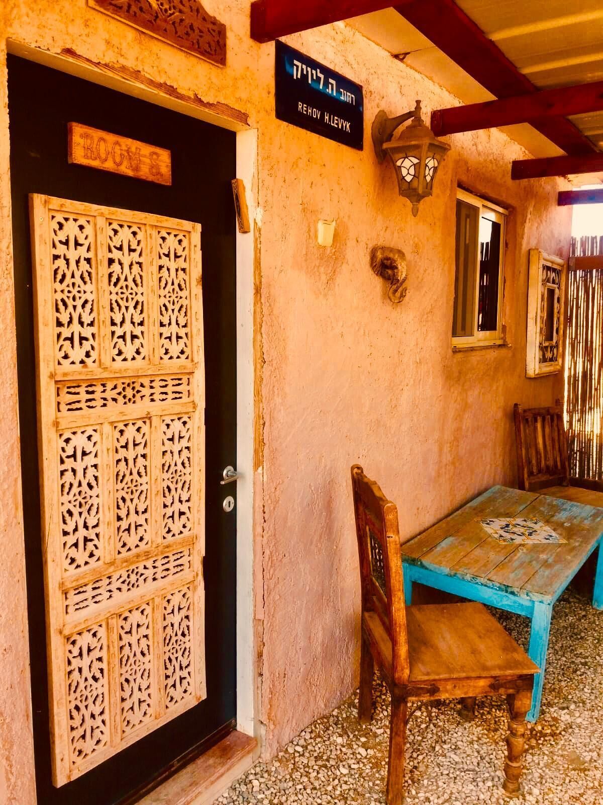 A wooden door with intricate carvings, a sign above it, and a table with a chair outside a building with a textured wall.