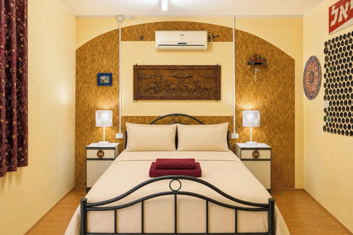 Habitación de hotel con cama, dos mesitas de noche, aire acondicionado de pared, un adorno en la pared y cortina estampada.