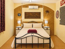 A hotel room with a bed, two bedside tables, a wall-mounted air conditioner, a decorative wall piece, and a patterned curtain.