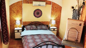 A bedroom with a bed, two bedside tables, a wall-mounted air conditioner, a decorative wall hanging, and a small plant.