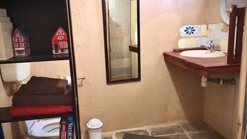 Un baño pequeño con inodoro, lavamanos, espejo y una estantería con libros y una maqueta de una casa.