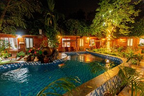 A pool area with wooden cabins, surrounded by lush greenery and illuminated by string lights.