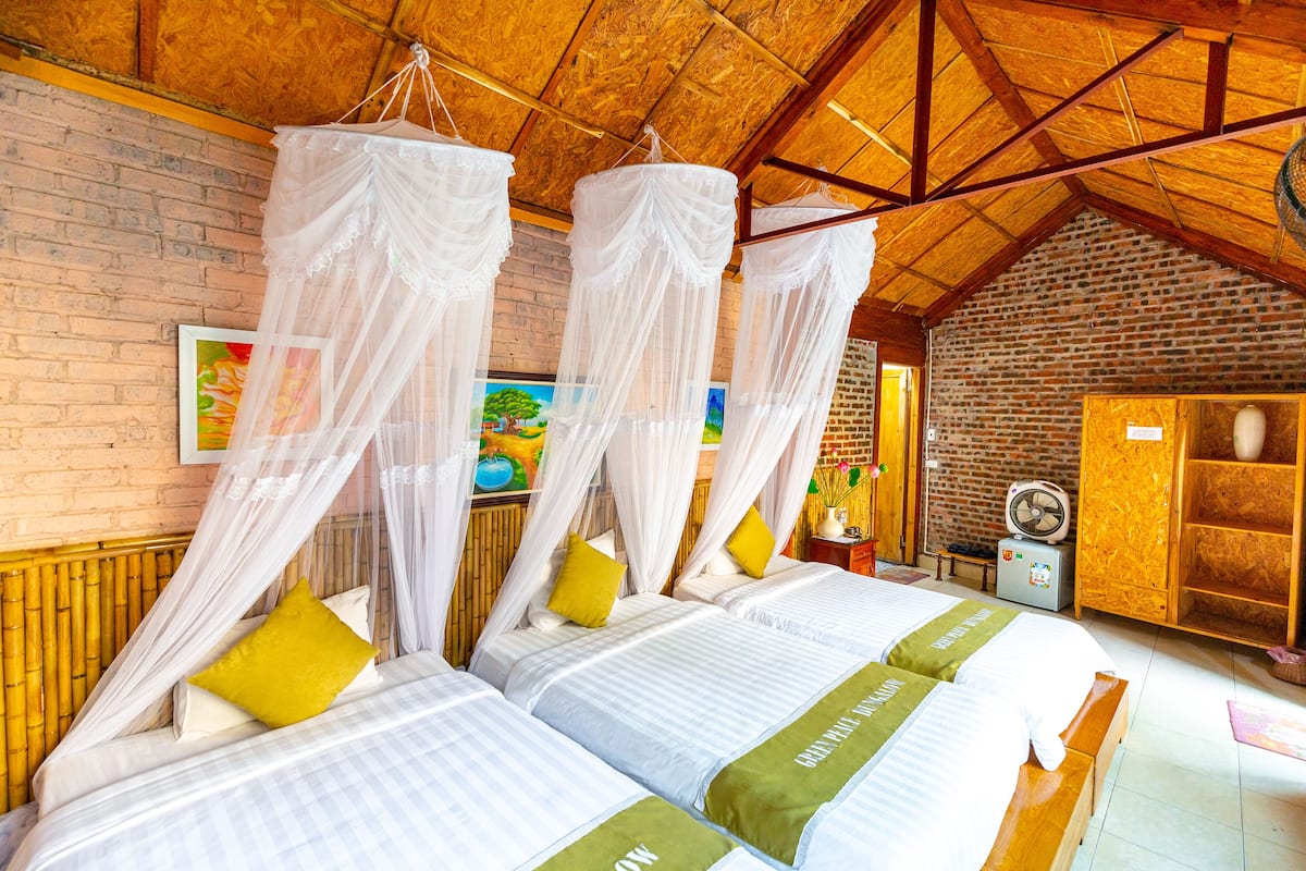 A room with two beds, each with a canopy bedspread and a green bed runner. There is a wooden bench and a small table with a fan.