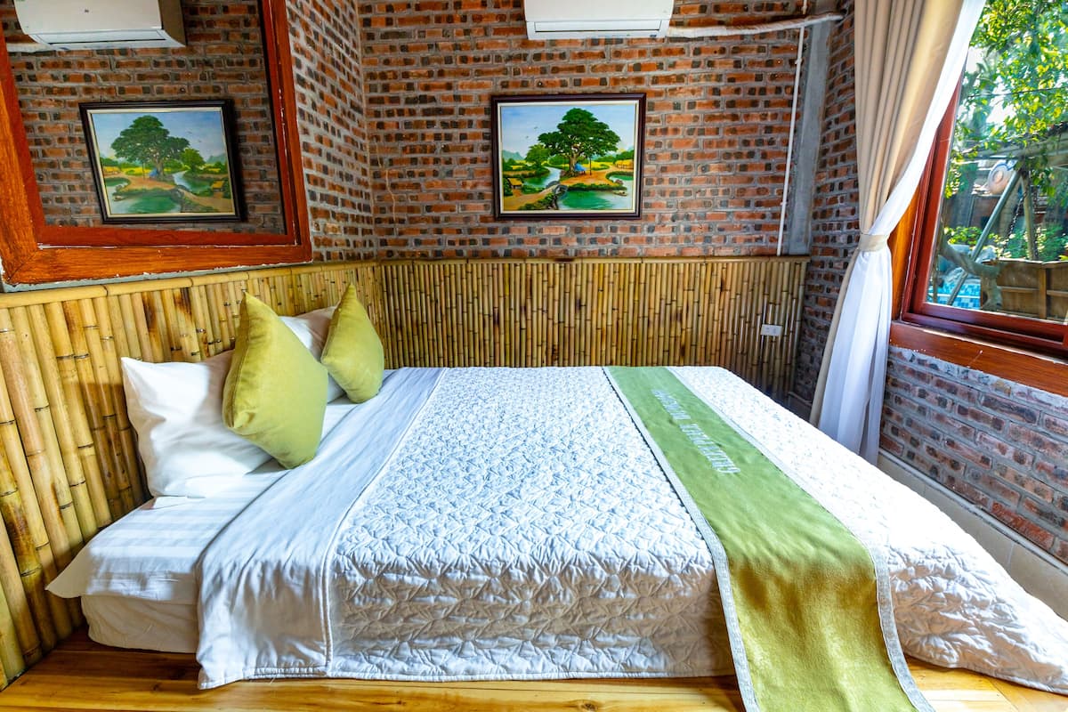 A room with a bed, bamboo wall paneling, a brick wall with two framed paintings, and a window with a view of greenery.