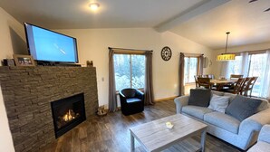 A living room with a stone fireplace, a television on a mantel, a grey sofa, a wooden coffee table, and a dining area with a chandelier. - Dogwood Dream @ Spring Brook Resort (Wisconsin Dells)
