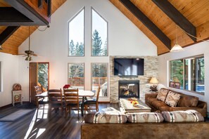 A spacious living room with a fireplace, large windows, and a high ceiling with exposed beams. - North Shore Cabin with Private Hot Tub (Kings Beach)