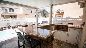 An outdoor kitchen with a stone countertop, bar stools, and a poolside view.