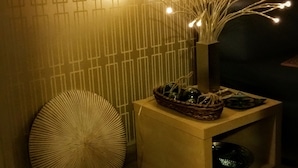 Ein Teil eines Raumes mit einer gemusterten Wand, einem kleinen Holfnachtkästchen mit einem Korb und einer Vase mit Lampen, sowie ein runder gewebter Hocker.