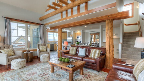 A spacious living room with a large brown leather sofa, a wooden coffee table, and a patterned rug. There is a staircase with wooden railings leading to an upper level. - Mountain Views, Steps from the Lifts (Big Sky)