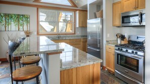 A modern kitchen with granite countertops, stainless steel appliances, wooden cabinets, and a large island with bar stools. - Mountain Views, Steps from the Lifts (Big Sky)