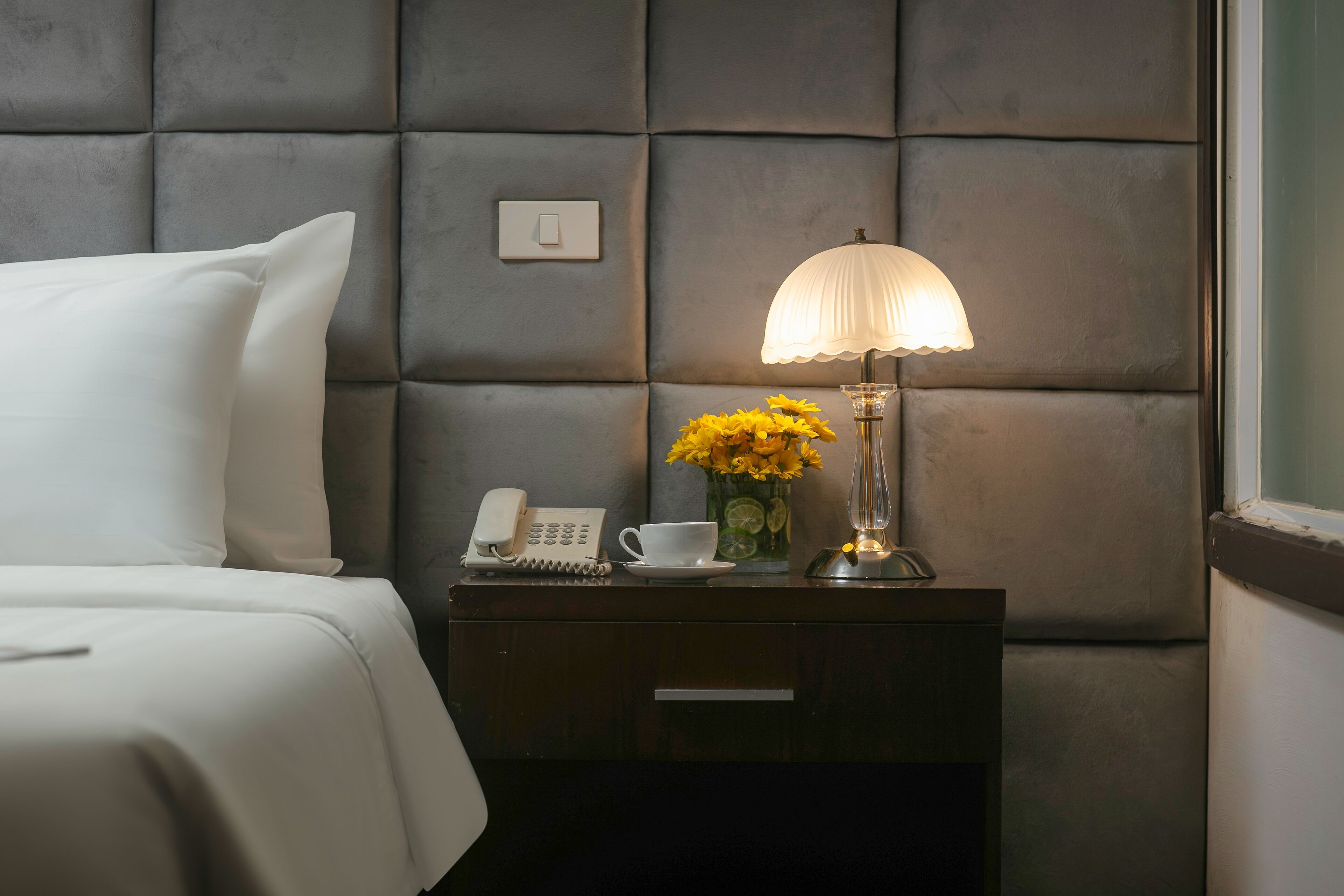 A neatly made bed with white linens, a bedside table with a lamp, a phone, a cup, and a vase with yellow flowers.