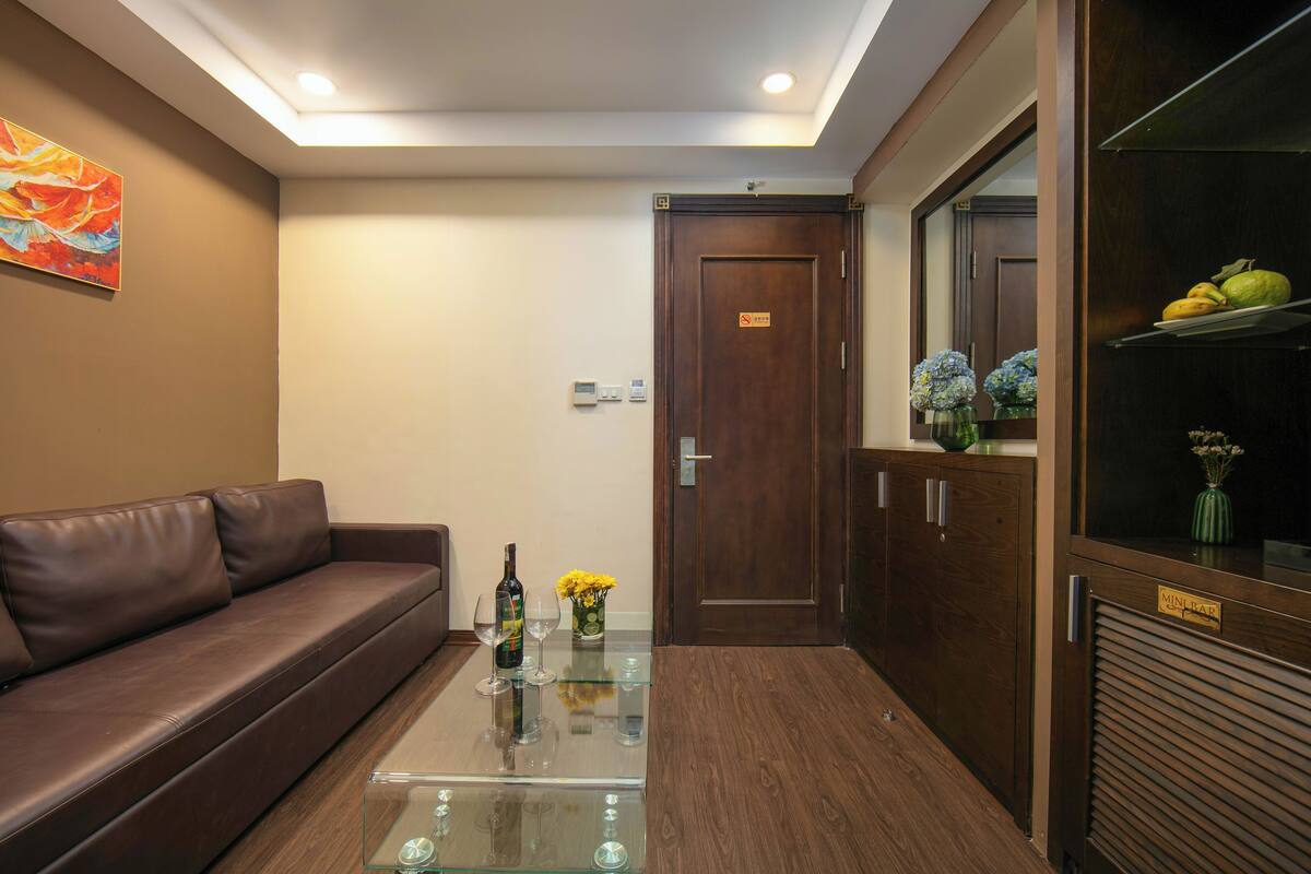 A reception area with a brown leather sofa, a glass coffee table with wine glasses, a wooden cabinet with a fruit display, and a door with a sign.
