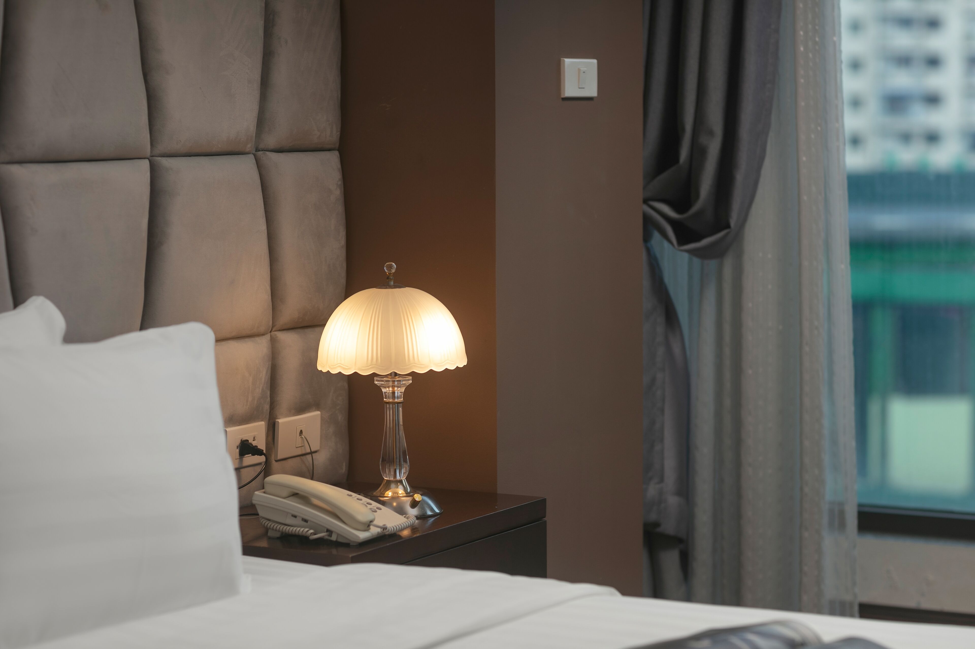 A hotel room with a bed, a bedside lamp, a telephone, and a window with curtains.