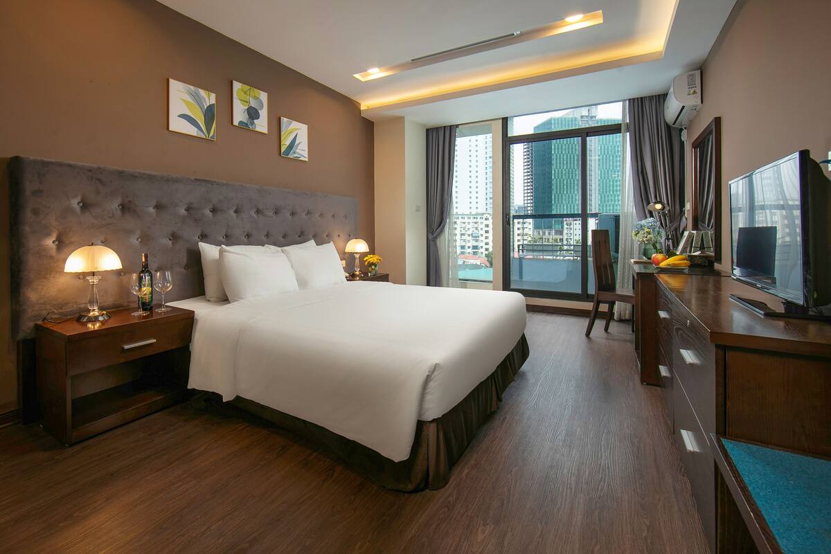 A hotel room with a large bed, a desk with a television, and a view of the city through large windows.