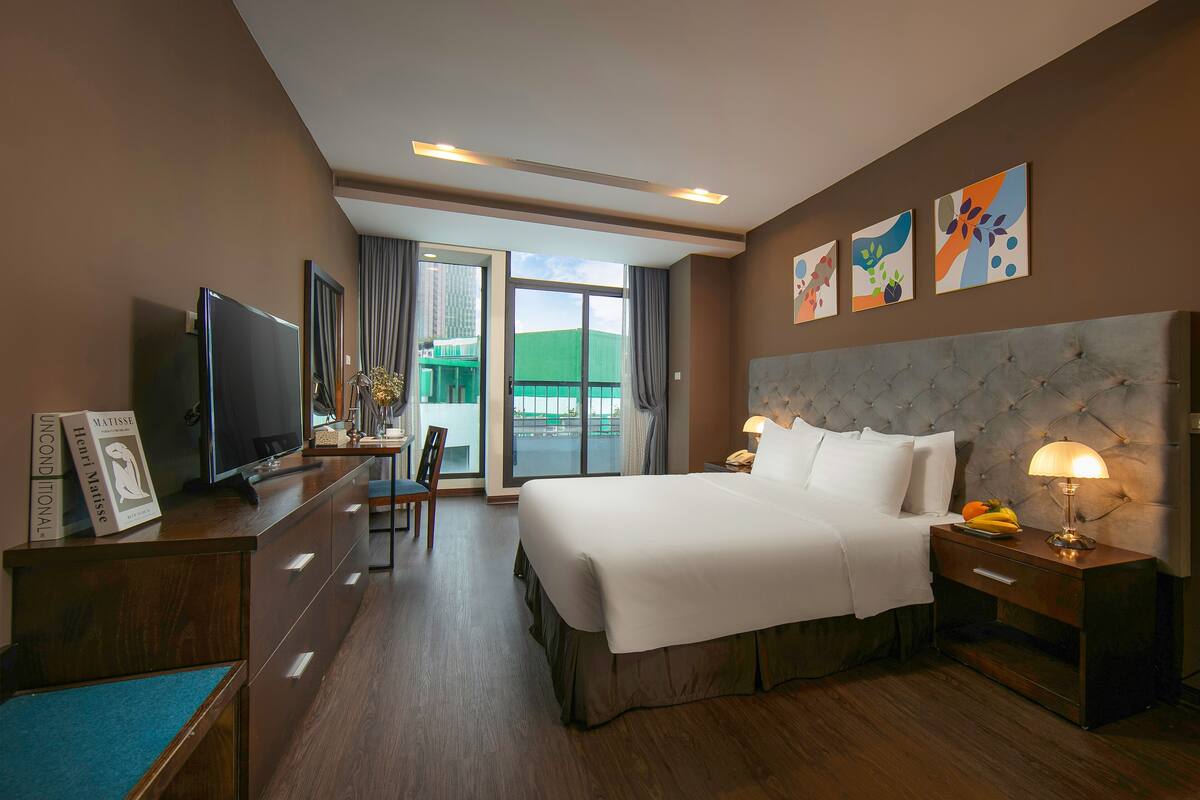 A hotel room with a large bed, a television, a desk with a computer, a chair, and a view of the outside.