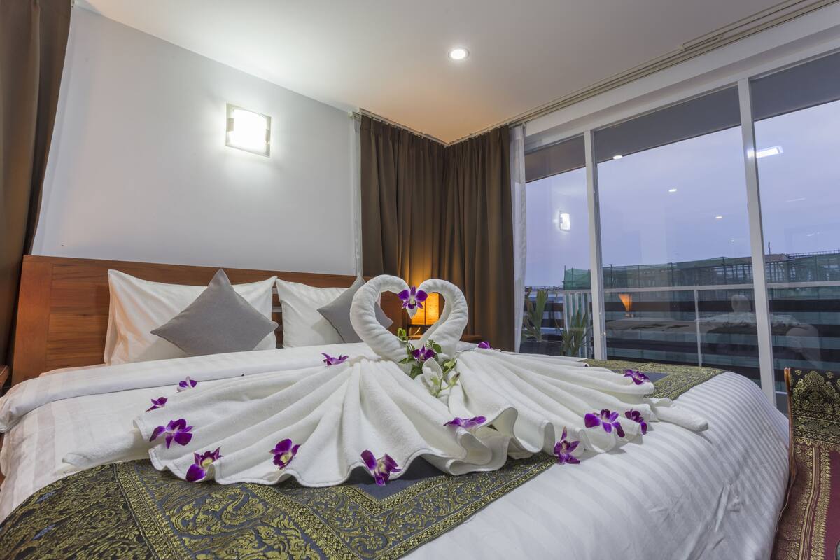 A hotel room with a large bed, a heart-shaped towel arrangement, and a view of the cityscape through the window.