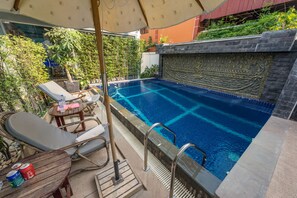 En swimmingpool med liggestole og et bord, omgivet af planter og en dekorativ væg.