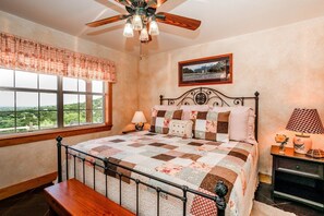 A bedroom with a large window, a bed with a quilt, a ceiling fan, a bedside table with a lamp, and a framed picture on the wall. - Deer Ridge Cottage | Pet Friendly Country Property (Fredricksburg)