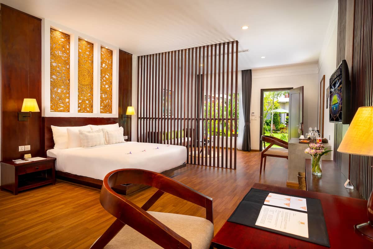 A hotel room with a large bed, wooden floor, a desk with a chair, a TV, and a decorative partition with a view of the garden.