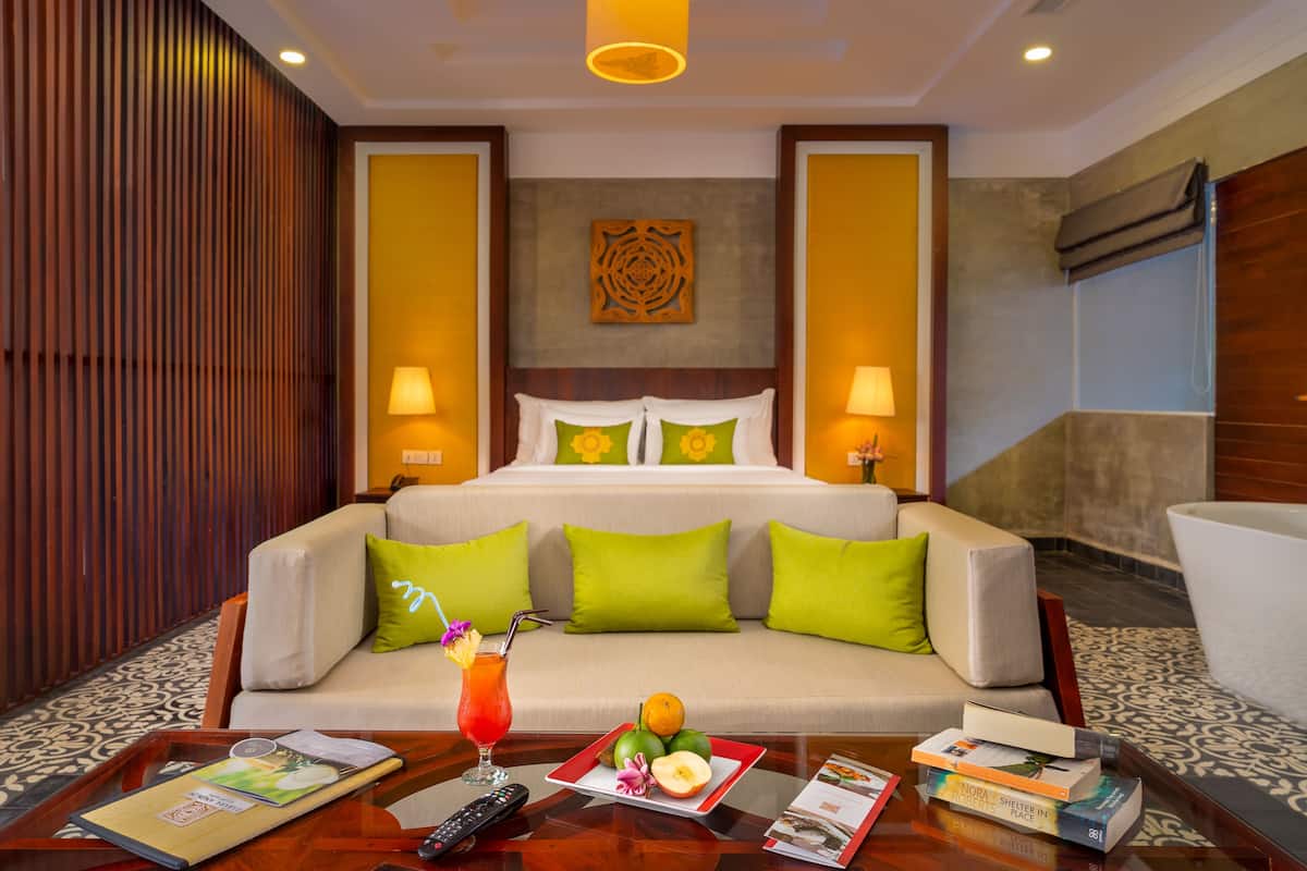 A hotel room with a bed, two bedside lamps, a wooden panel wall, a decorative wall piece, a sofa with green cushions, a coffee table with a tray, a drink, fruit, and books.