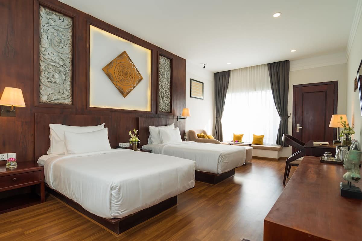 A hotel room with two beds, wooden flooring, decorative wall panels, a desk with a chair, and a window with curtains.