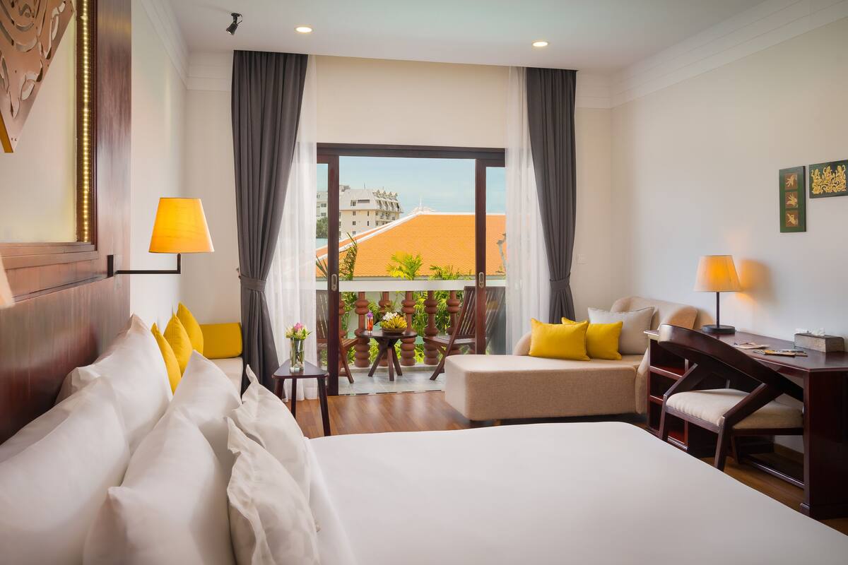 A hotel room with a large bed, a desk, a chair, a balcony with a view of buildings and greenery.