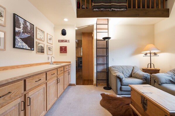 A cozy living room with a sofa, a wooden cabinet, and a lamp. There is a ski lift sign on the wall and a framed picture of a snowy landscape. - Walk to Dining, Shops | Hot Tub | Sauna (Ketchum)