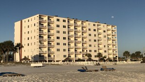 Un hotel de varios pisos con balcones, cerca de una playa con tumbonas y palmeras.