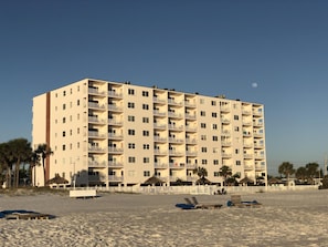 A multi-story hotel with balconies, located near a beach with sun loungers and palm trees.