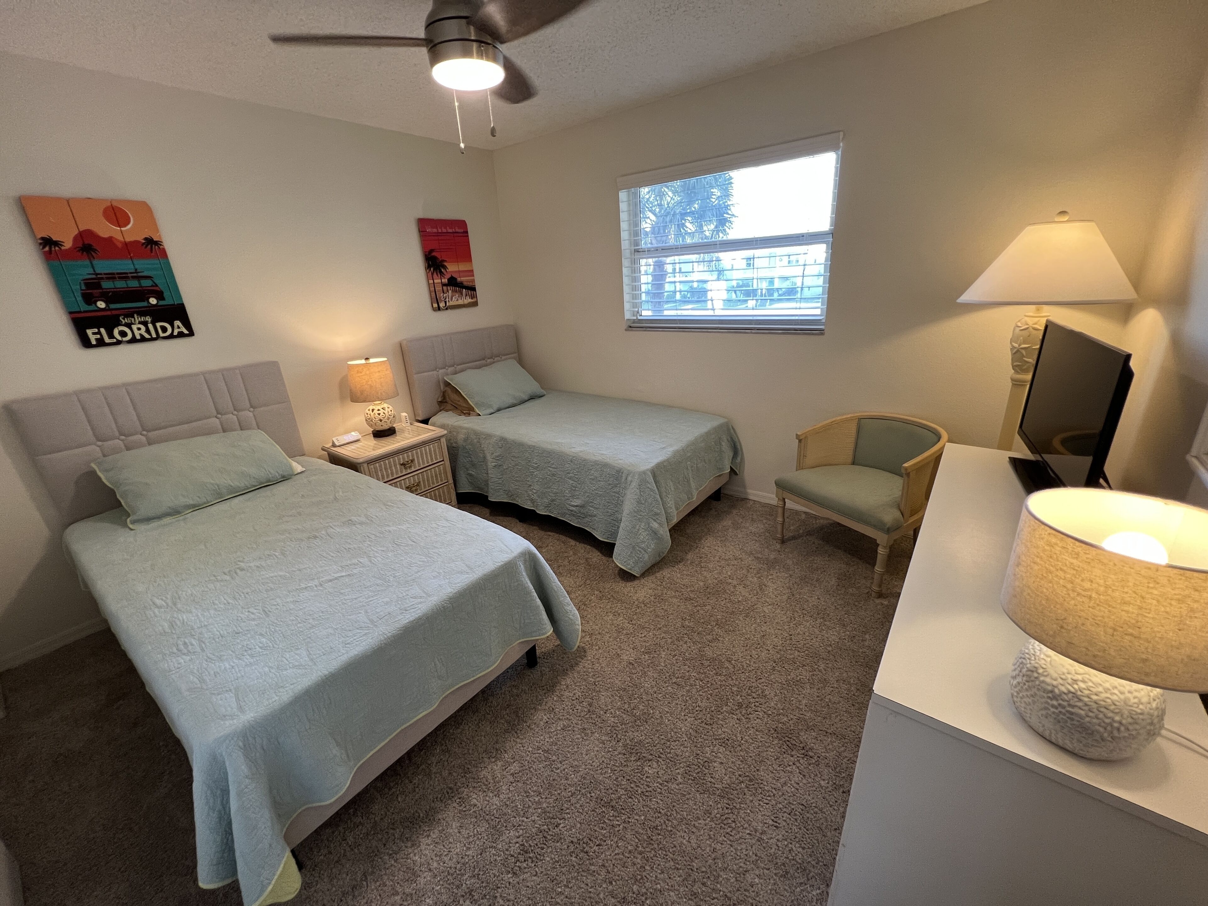 A bedroom with two beds, a chair, a desk with a lamp, a ceiling fan, and a window with a view of buildings.