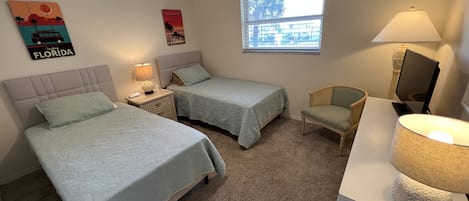 A bedroom with two beds, a chair, a desk with a lamp, a ceiling fan, and a window with a view of buildings.